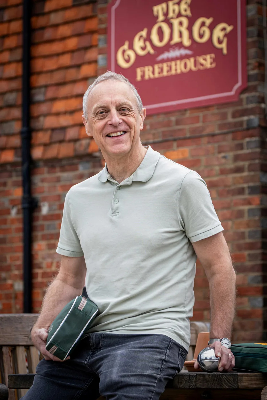 Colin Clifton, chair of The George Ltd, sitting on a bench outside The George with a boule in his hand and the pub's Freehouse sign behind him