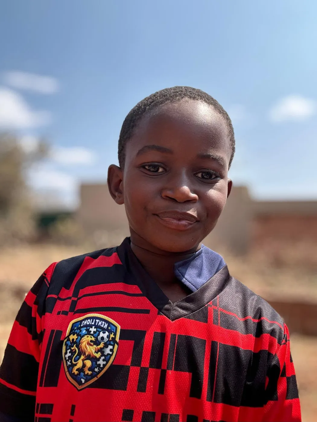 A smiling boy at Chidobe Primary School in Zimbabwe wearing a red and black Kent Lions football shirt with the club's lion badge