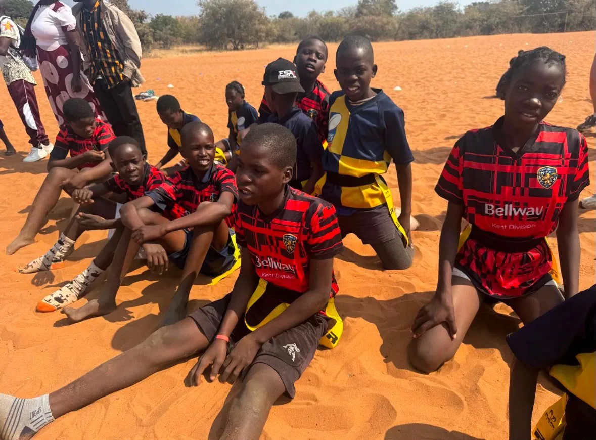 A group of pupils sitting and kneeling on red sand, several wearing red and black Kent Lions shirts with the Bellway Kent Division sponsor name, others in navy and yellow tag rugby tops