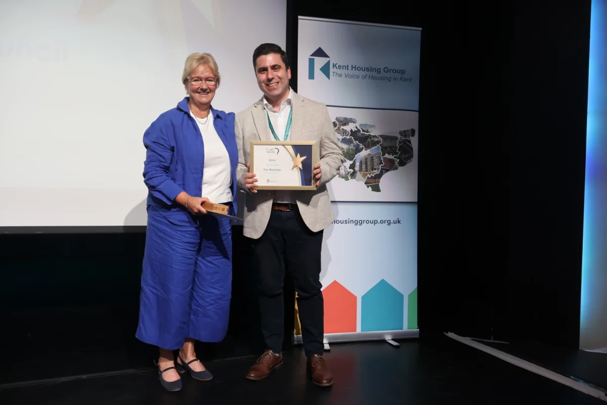Ashford Borough Council chief executive Tracey Kerly presenting the Kent Housing Group Chair's Award certificate to Tim Woolmer of Kent County Council on stage in front of a Kent Housing Group banner