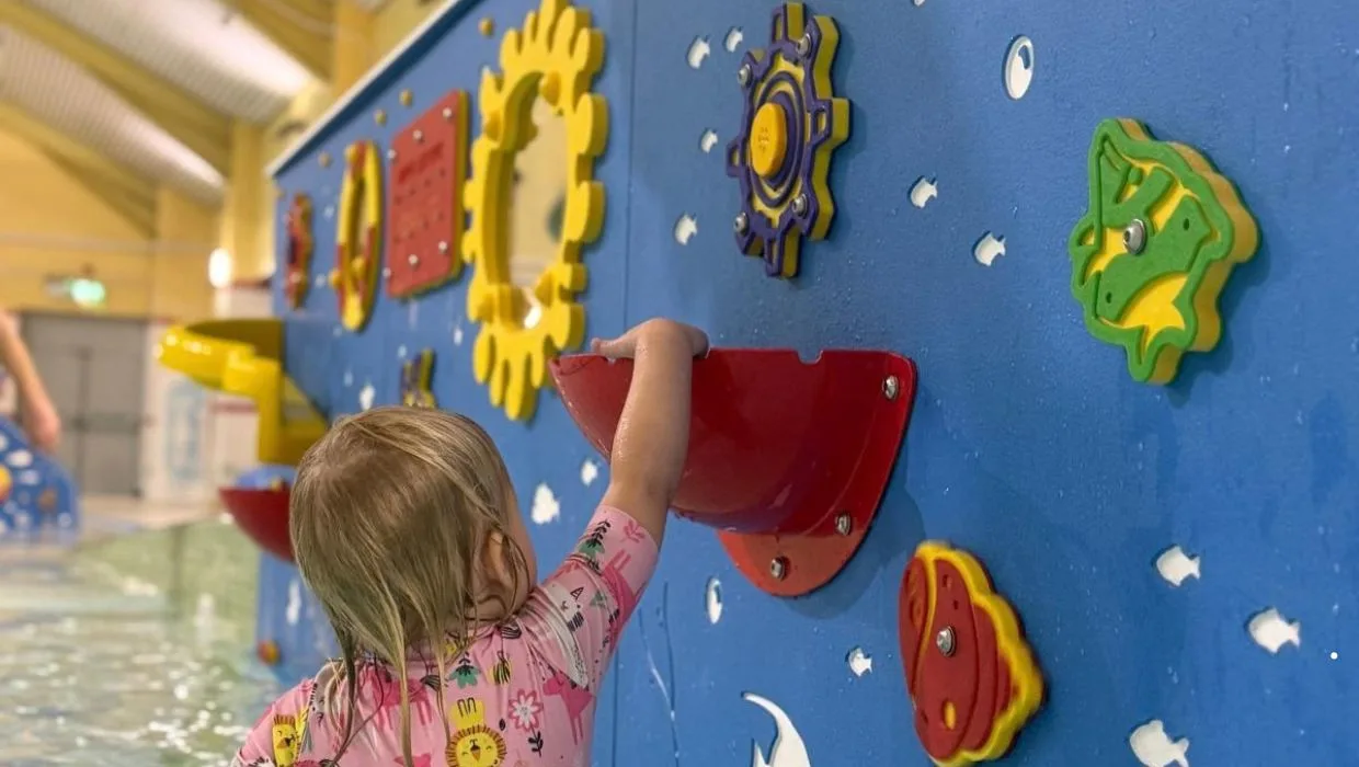A young child in a swimsuit reaching up to a red water bucket on a blue water-play wall beside the pool at Tenterden Leisure Centre